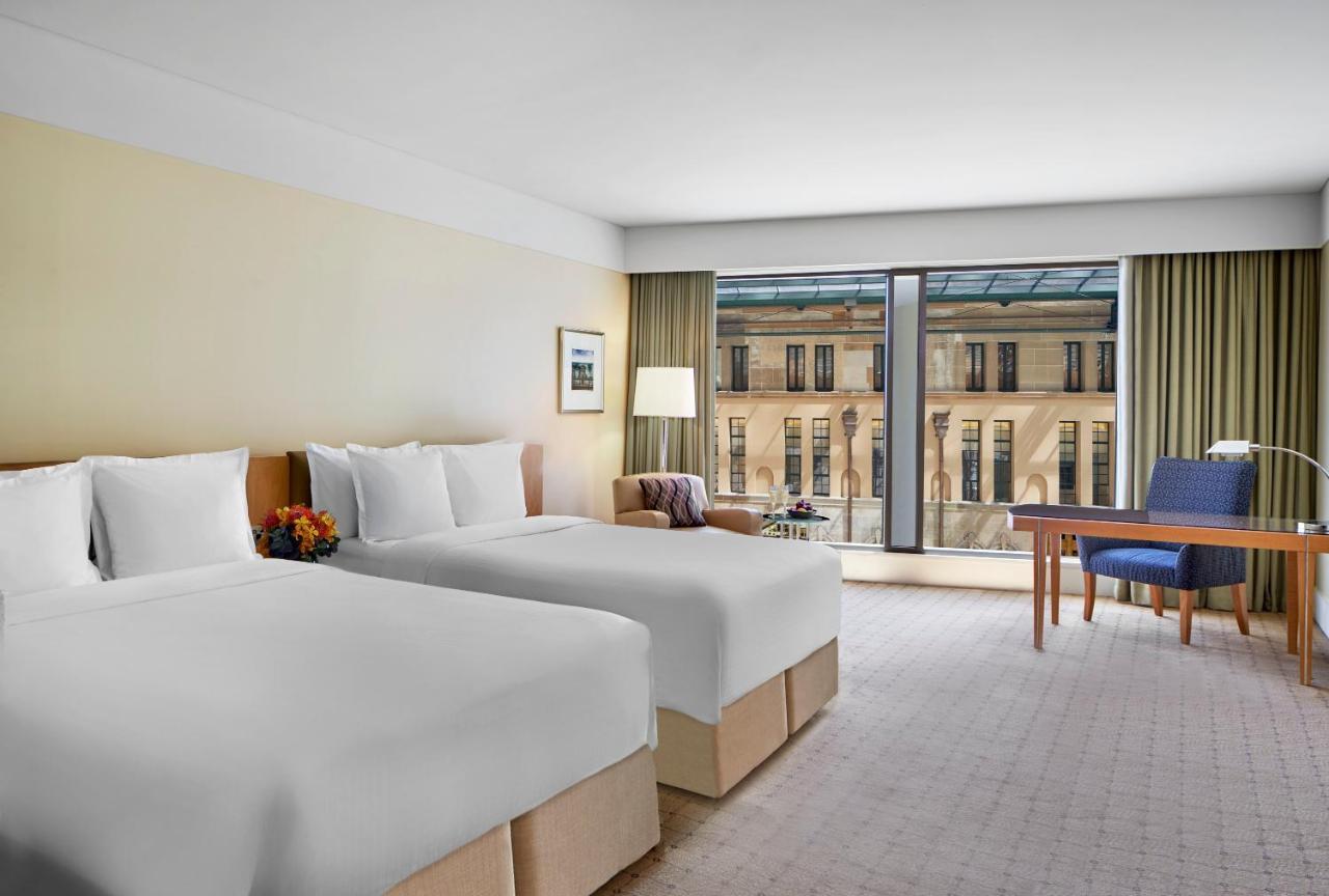 The Fullerton Hotel Sydney Exterior foto The photo shows a modern hotel room with two neatly made beds, each adorned with white bedding and pillows. There is a bedside table with a small decorative arrangement and a lamp beside one of the beds. The room features large windows with sheer cur