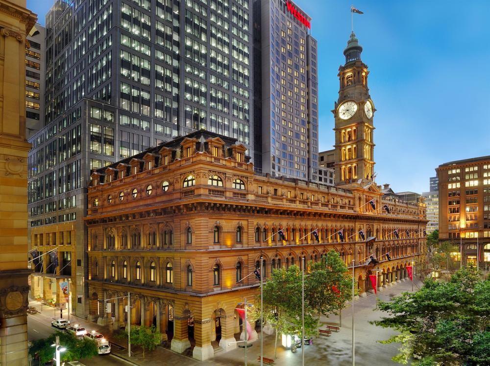 The Fullerton Hotel Sydney Exterior foto The image depicts a contrasting architectural scene featuring a historic building alongside modern skyscrapers. The foreground shows a grand, ornate structure with decorative elements, likely a former post office or civic building, distinguished by i