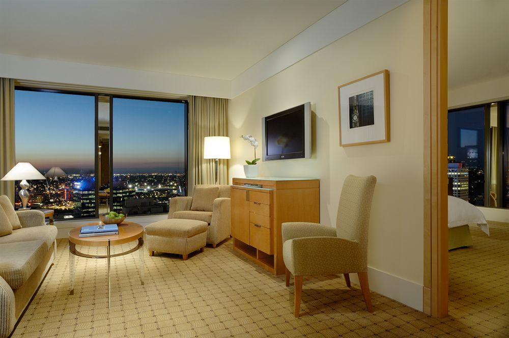 The Fullerton Hotel Sydney Exterior foto The photo shows a hotel room with a modern and elegant design. There's a large window with a view of a city skyline, indicating it's likely at a higher floor. The room features a seating area with two light-colored chairs and a small round coffee tab