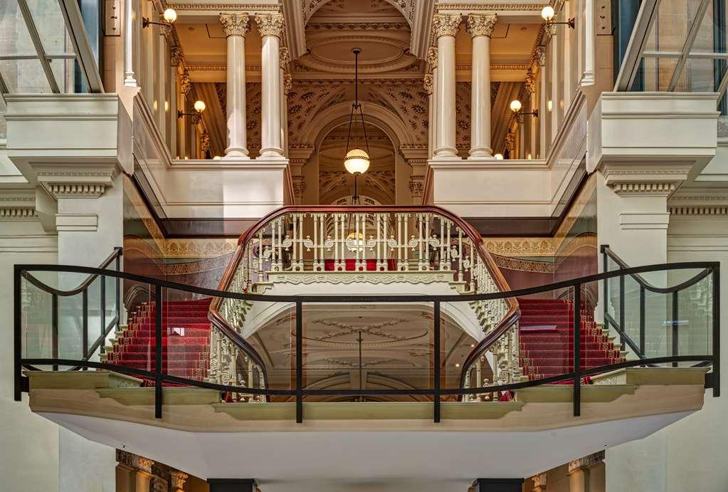 The Fullerton Hotel Sydney Interior foto The photograph depicts an elegant interior of a building, showcasing a grand staircase that curves gracefully between floors. The staircase features intricate railings and a plush red carpet that adds a touch of luxury. The space is adorned with tall