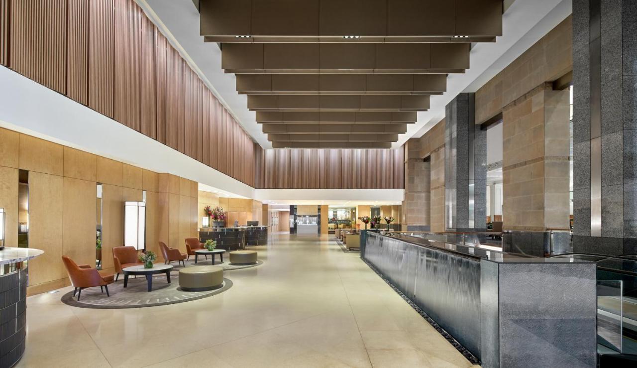 The Fullerton Hotel Sydney Exterior foto The photo shows a spacious and modern hotel lobby. It features high ceilings with large, rectangular light fixtures hanging above. The walls are a combination of warm wood tones and stone, creating an elegant atmosphere. On one side, there are comfor
