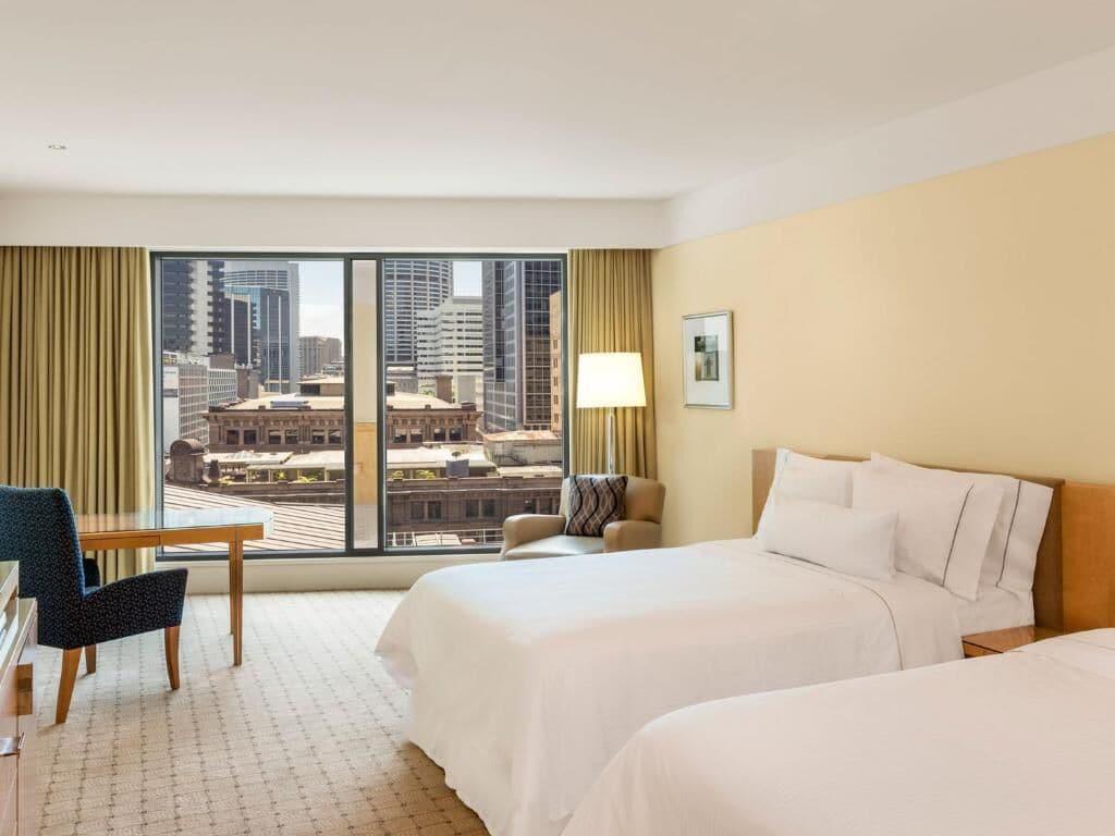 The Fullerton Hotel Sydney Exterior foto The photo shows a hotel room with a modern design. There are two beds with white linens positioned in the center. A large window offers a view of a cityscape, featuring tall buildings outside. To the side, there is a comfortable armchair and a wooden