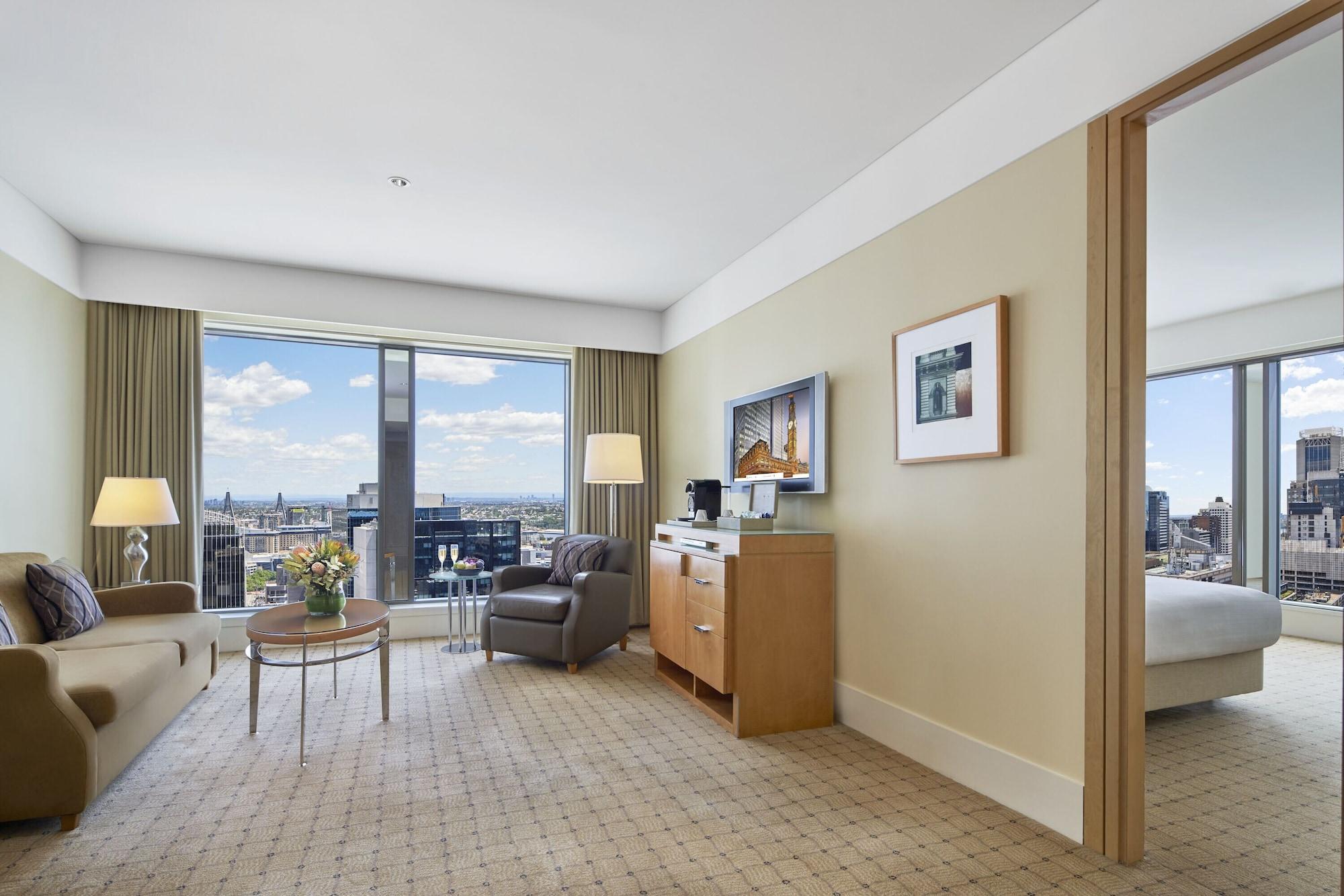 The Fullerton Hotel Sydney Exterior foto The photo shows the interior of a hotel room. It features a spacious living area with large windows that offer a view of the city skyline. The room has beige walls and carpet, giving it a warm ambiance. There is a comfortable sofa, a coffee table, an