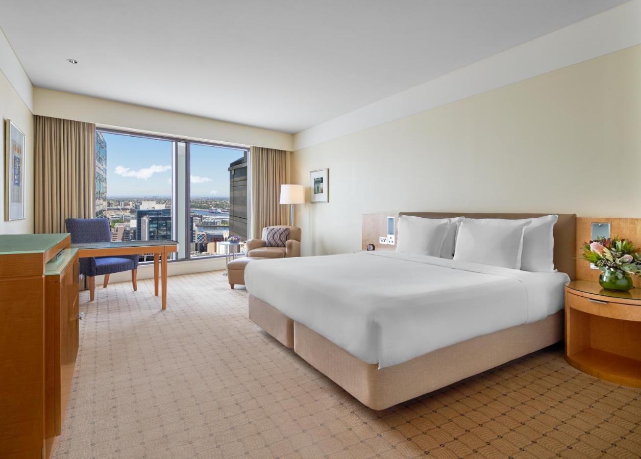 The Fullerton Hotel Sydney Exterior foto The photo shows a hotel room featuring a large bed with white linens, set in a spacious area with light-colored walls. To one side, there is a comfortable chair and a small table, both positioned near a large window that allows for natural light and 