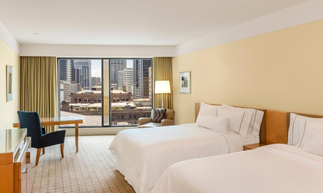 The Fullerton Hotel Sydney Exterior foto The photo shows a hotel room featuring two beds with white bedding. The walls are painted a soft yellow, creating a warm atmosphere. Large windows provide a view of the city skyline, and there are beige curtains framing the windows. In the room, ther