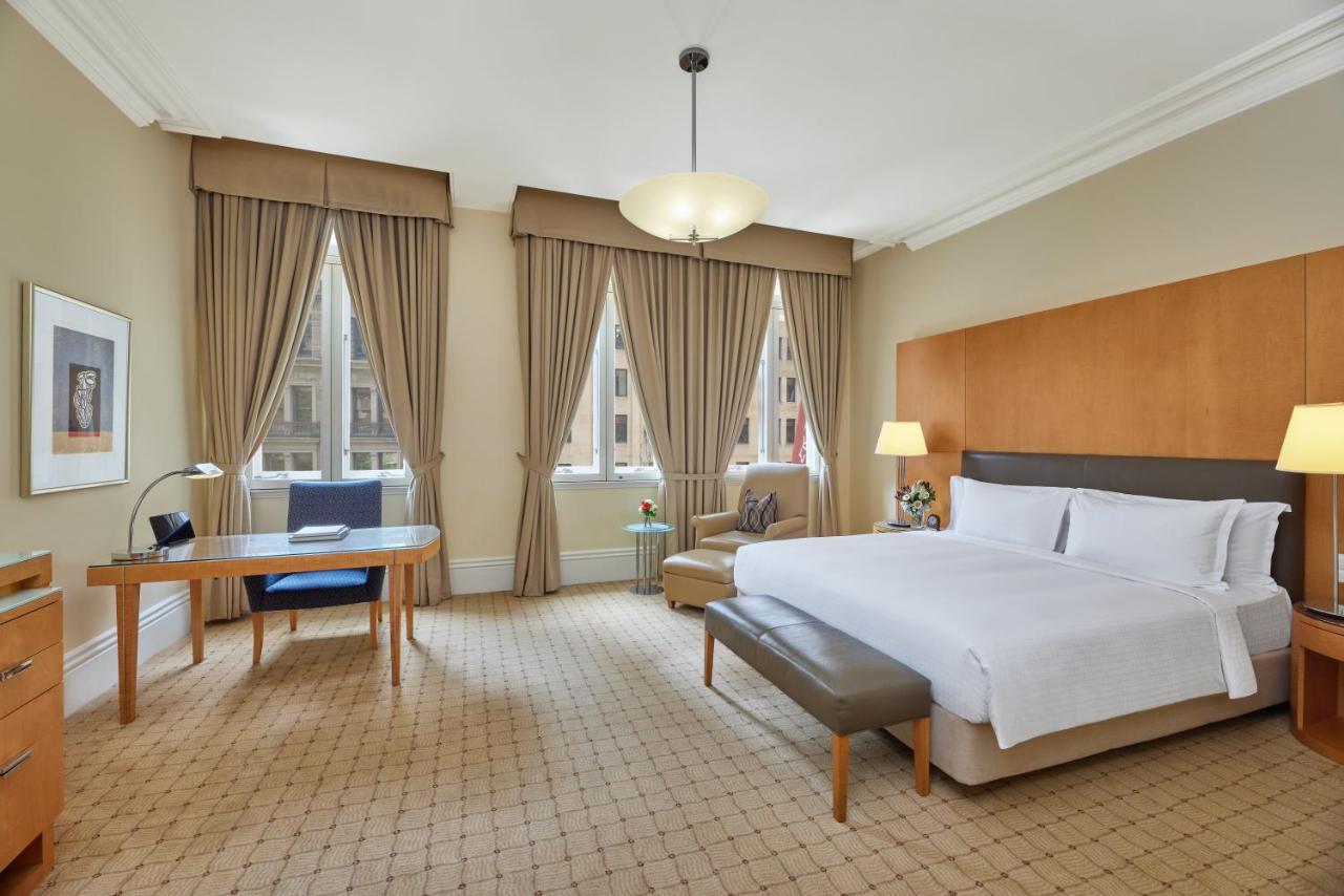 The Fullerton Hotel Sydney Exterior foto The photo shows a spacious hotel room featuring a large bed with white linens. There is a modern bedside table and a small couch at the foot of the bed. A comfortable armchair is positioned nearby. Natural light enters through large windows adorned w