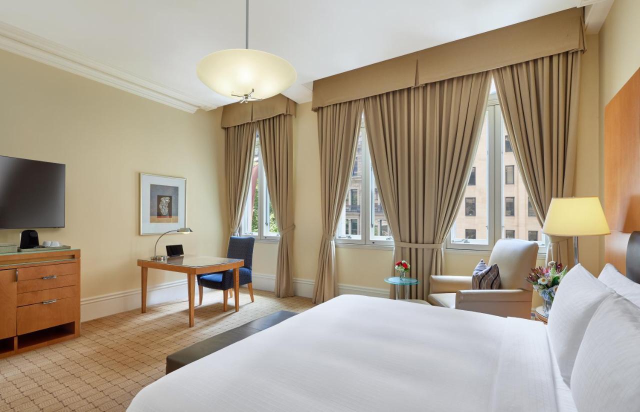 The Fullerton Hotel Sydney Exterior foto The photo shows a well-furnished hotel room. It features a large bed with white linens, a desk with a chair, and a comfortable armchair beside it. The room has large windows draped with beige curtains, allowing natural light to enter. A flat-screen T