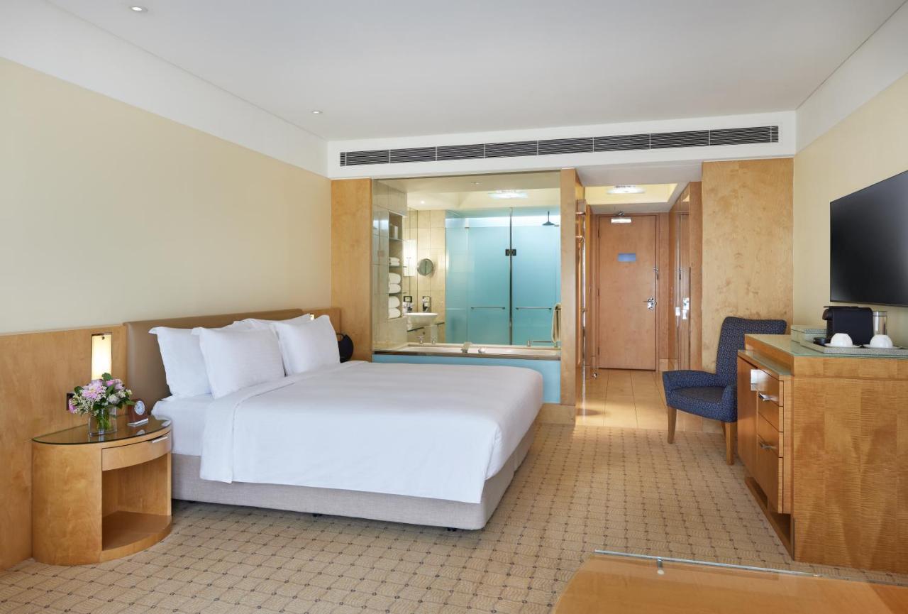 The Fullerton Hotel Sydney Exterior foto The photo shows a modern hotel room featuring a spacious layout. It includes a large bed with white linens, a wooden bedside table with a flower arrangement, and a chair in the corner. The room has a light-colored, neutral palette, giving it a clean 