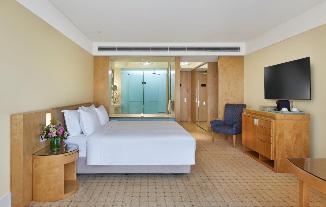 The Fullerton Hotel Sydney Exterior foto The photo shows a modern hotel room. It features a large bed with white linens, a small bedside table with a vase of flowers, and a comfortable blue armchair in one corner. There is a wooden dresser and a flat-screen TV mounted on the wall. Behind th