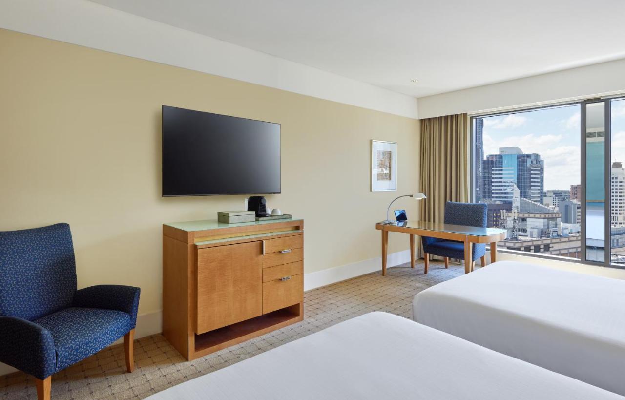 The Fullerton Hotel Sydney Exterior foto The photo shows a modern hotel room featuring two beds with white linens. There's a wooden bedside table and a blue upholstered chair nearby. A television mounted on the wall is visible, alongside a wooden dresser with some items on top. Additionally