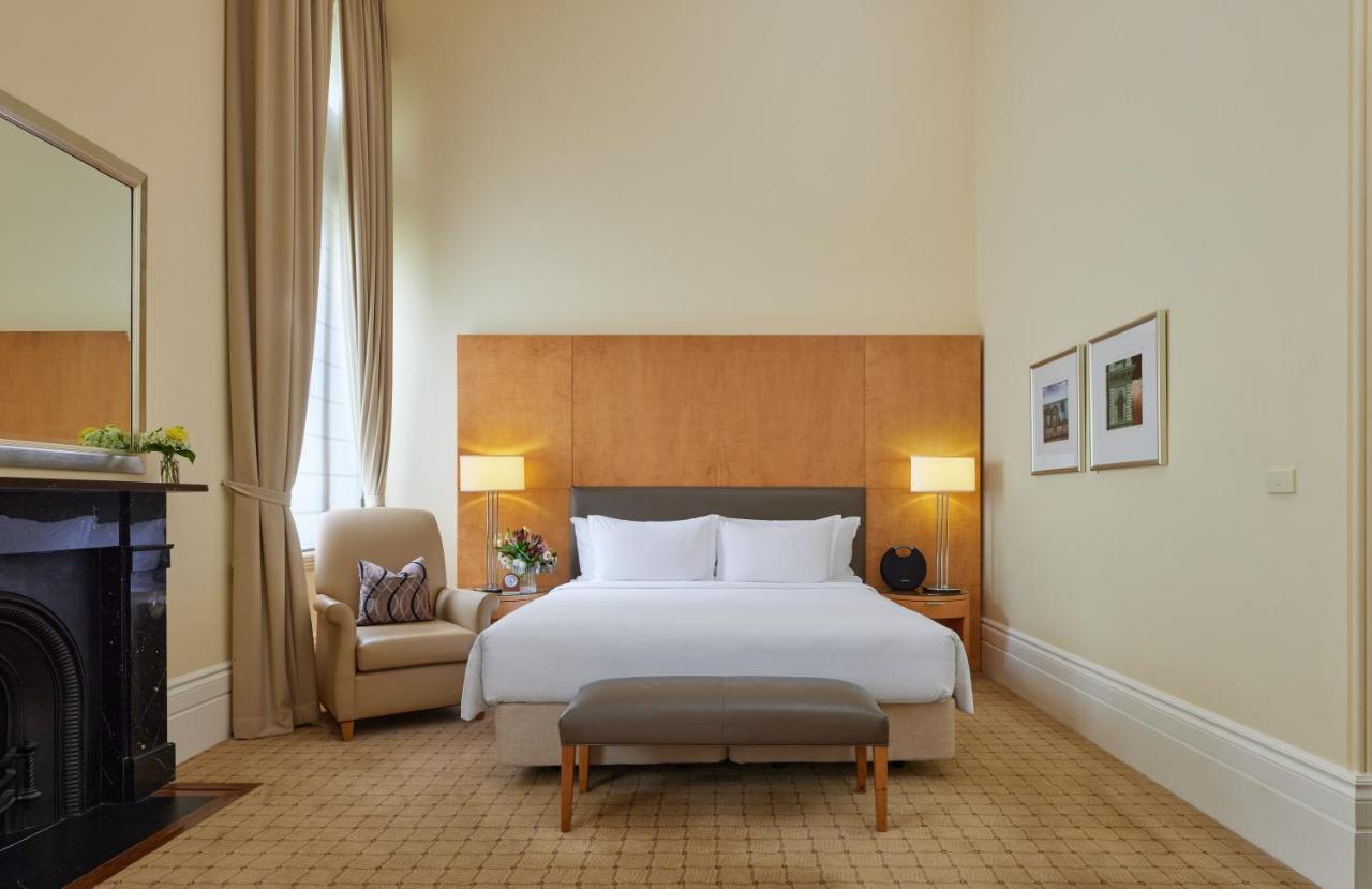The Fullerton Hotel Sydney Exterior foto The photo shows a well-appointed hotel room. It features a large bed with white linens and a gray headboard, positioned against a wooden wall panel. On either side of the bed are stylish lamps. There is a comfortable armchair in the corner, accompani
