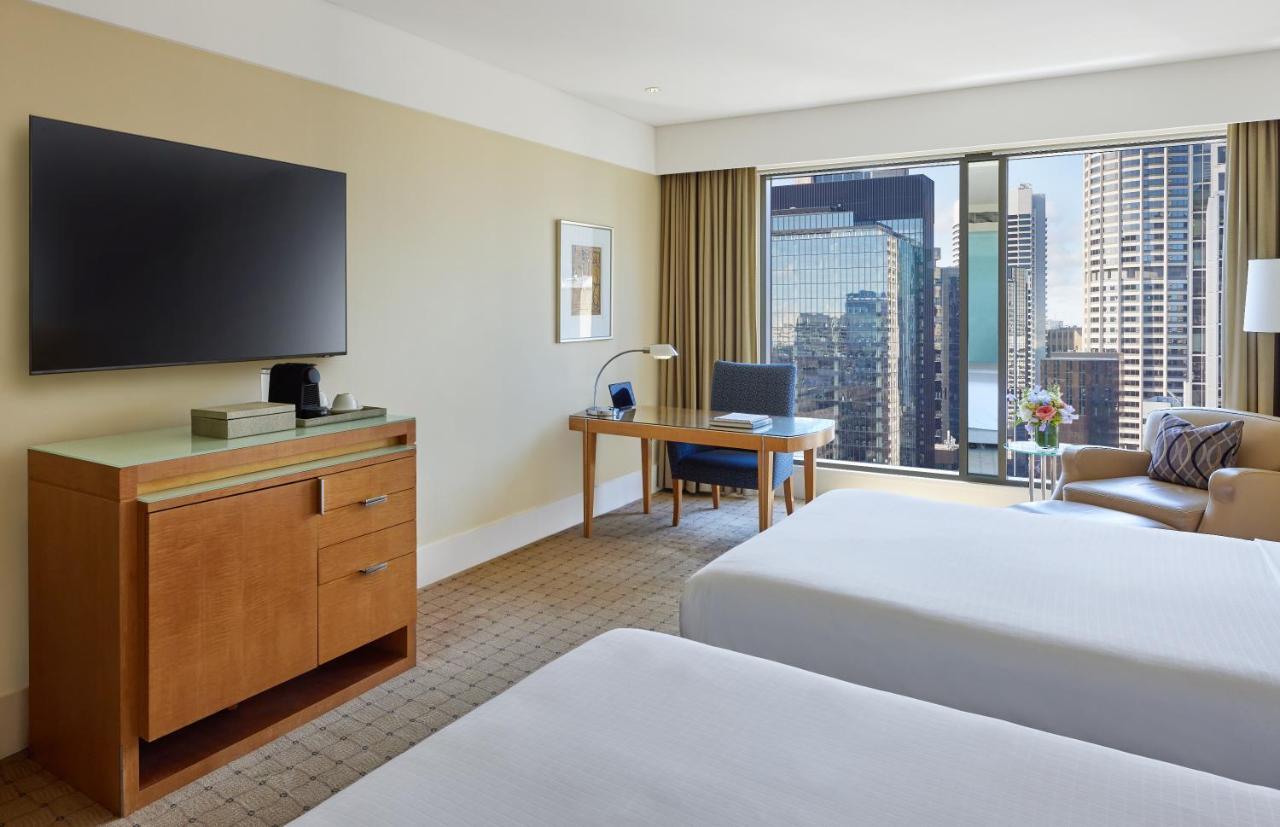 The Fullerton Hotel Sydney Exterior foto The photo shows a hotel room featuring two beds with white bedding. There is a wooden furniture piece with a television mounted on the wall above it. To the side, there is a desk with a chair and a lamp, positioned near a large window that offers a v
