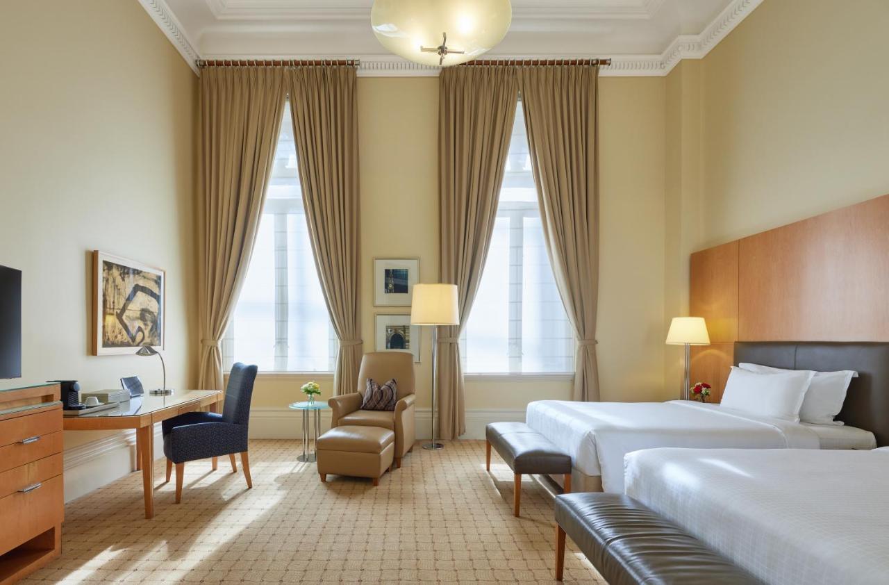 The Fullerton Hotel Sydney Exterior foto The photo shows a well-appointed hotel room featuring two beds. The room has high ceilings and large windows dressed with elegant drapes that allow natural light to fill the space. There is a stylish desk and chair positioned against one wall, alongs