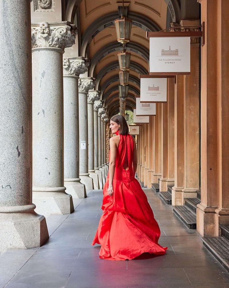 The Fullerton Hotel Sydney Exterior foto The photo depicts a woman wearing an elegant red gown, standing in a covered archway lined with columns. She is turned slightly to look back over her shoulder, showcasing the flow and design of the dress. The environment features multiple arches and 