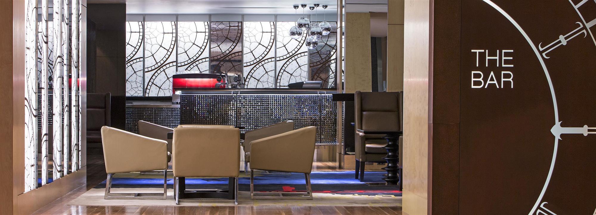 The Fullerton Hotel Sydney Exterior foto The photo shows a stylish interior of a hotel lobby or bar area. In the foreground, there are several beige lounge chairs arranged around a low table, creating a cozy seating area. The background features a modern bar with a sleek, dark counter and a